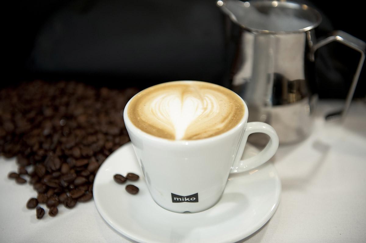 A white coffee cup labeled ‘miko’ with a heart design on the foam, placed on a black surface with coffee beans and a silver coffee pot in the background.